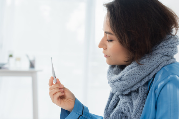 sick young woman in blue knitted scarf holding electronic thermometer - 写真・画像