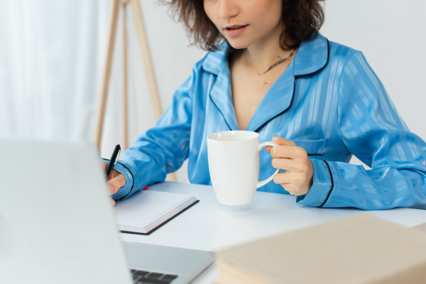 καλλιεργημένη άποψη της νεαρής γυναίκας κρατώντας φλιτζάνι καφέ κοντά σημειωματάριο και φορητό υπολογιστή - Φωτογραφία, εικόνα
