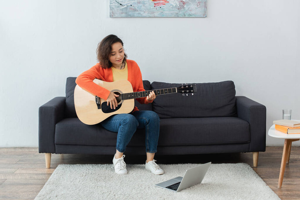 Ευτυχισμένη νεαρή γυναίκα μαθαίνει πώς να παίξει ακουστική κιθάρα κοντά στο φορητό υπολογιστή στο χαλί - Φωτογραφία, εικόνα
