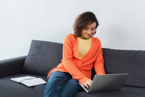 cheerful woman sitting on couch and using laptop near notebook - Photo, Image