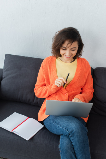 high angle view of cheerful woman sitting on couch and using laptop near notebook - Foto, afbeelding
