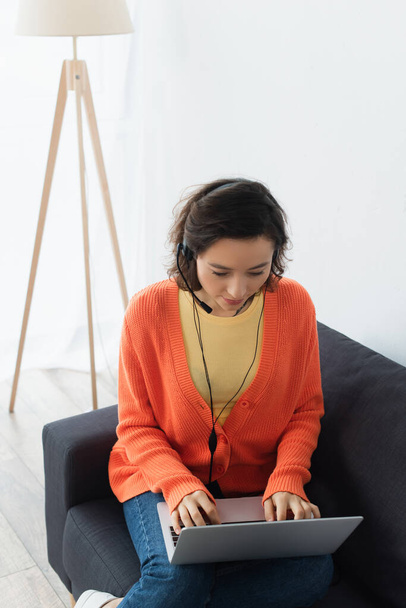 brunetka mladý operátor ve sluchátkách pomocí notebooku na gauči - Fotografie, Obrázek