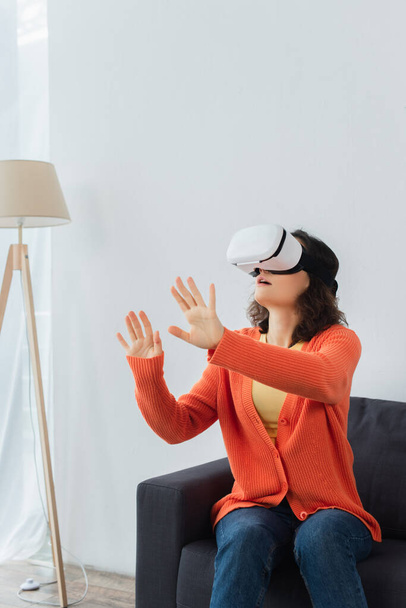 young brunette woman in vr headset gesturing while playing and sitting on sofa - Foto, Imagen