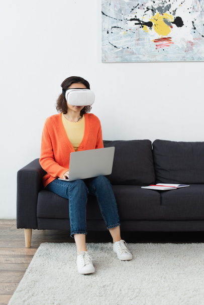 young brunette woman in vr headset using laptop while sitting on sofa  - 写真・画像
