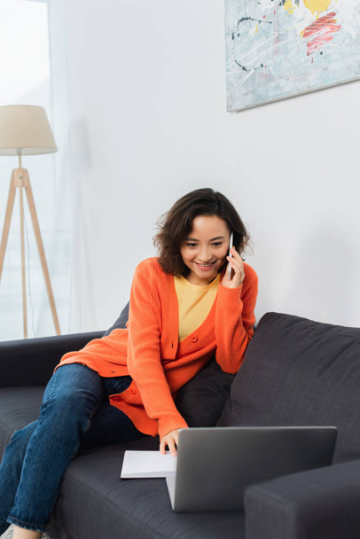 happy young woman talking on cellphone and using laptop on sofa  - Фото, зображення