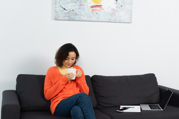 若い女性はソファの上に空白の画面を持つガジェットの近くにコーヒーのカップを保持 - 写真・画像