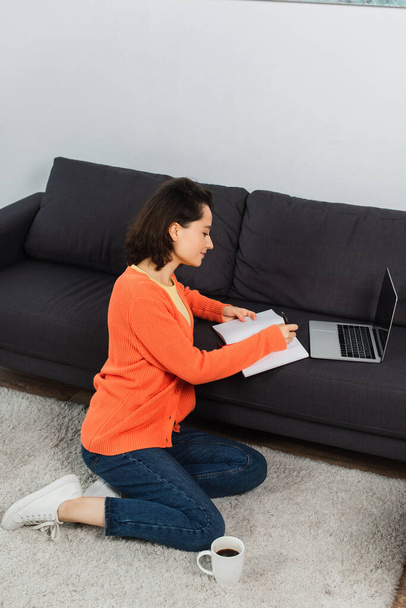 young woman sitting on carpet and writing in notebook near laptop with blank screen on sofa - Photo, Image