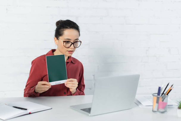 young teacher in eyeglasses holding card with green lettering and looking at laptop on desk  - Foto, imagen