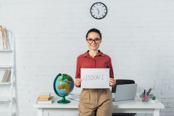 smiling woman in eyeglasses holding paper with lesson one lettering near globe and laptop on desk - Photo, Image
