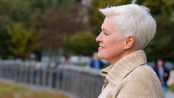 Closeup portrait 65s happy smiling mature lady in casual clothes standing posing outdoors turns head looking aside. Calm attractive benevolent woman of retirement age with gray stylish short hair - Photo, Image