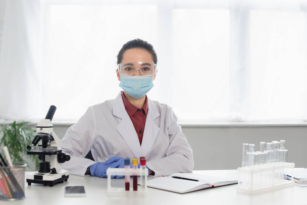 ученый в очках и медицинской маске смотрит на камеру возле пробирки и микроскопа - Фото, изображение