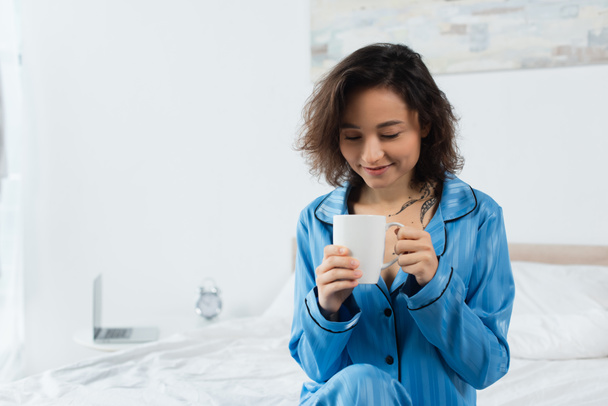 happy woman in blue pajamas holding cup of coffee in bedroom  - Zdjęcie, obraz