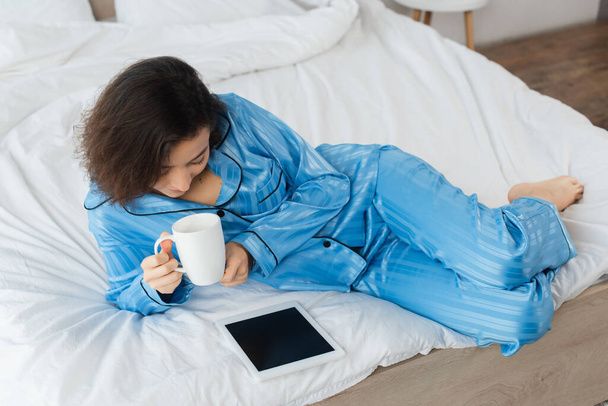 vista de ángulo alto de la mujer en pijama azul que sostiene la taza de café y el uso de la tableta digital en el dormitorio  - Foto, Imagen