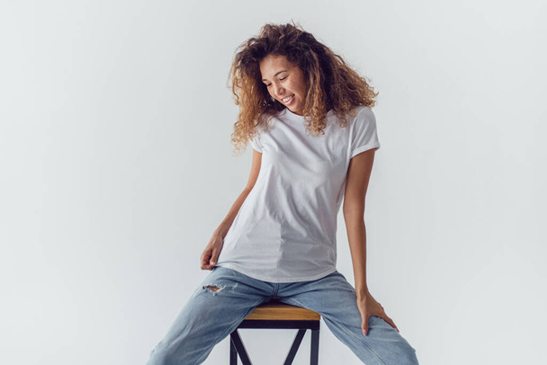Smiling woman with lush curly hair in white blank t-shirt sits on a white background. Mock-up. - Zdjęcie, obraz