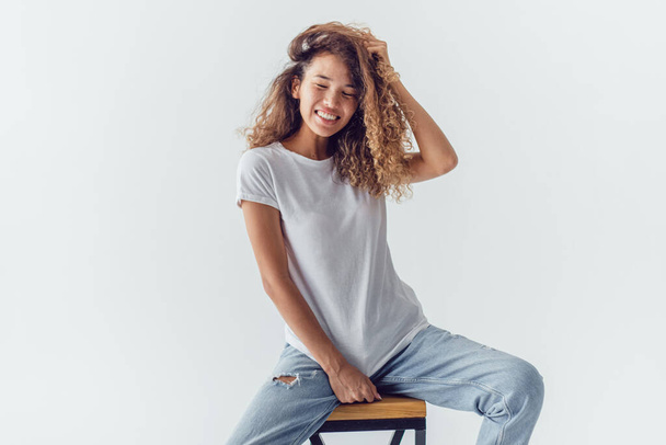 Smiling woman with lush curly hair in white blank t-shirt sits on a white background. Mock-up. - Foto, Bild