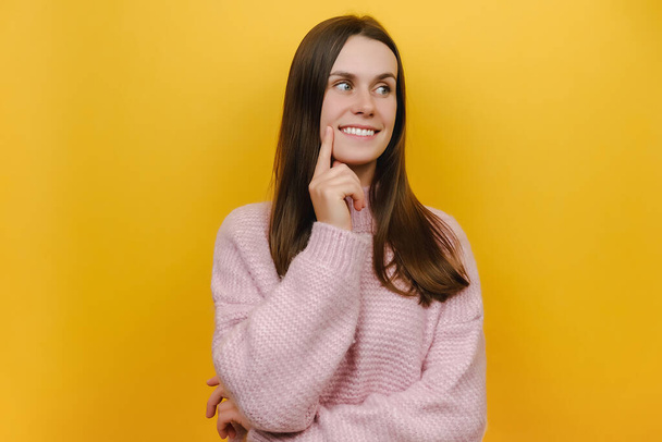 Portrait of cute playful young woman smiling and thinking wears pink knitted sweater, posing isolated over yellow color background wall with copy space for promotion content. People emotion concept - Valokuva, kuva