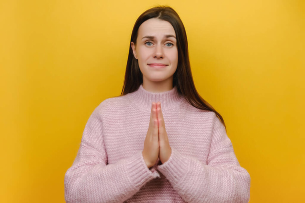 Close up portrait of pleaded begging asking sad young caucasian woman 20s hands folded in prayer gesture, begging about something, dressed in pink knitted sweater, isolated on yellow studio background - Foto, imagen