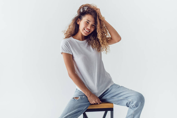 Smiling woman with lush curly hair in white blank t-shirt sits on a white background. Mock-up. - Foto, Imagem