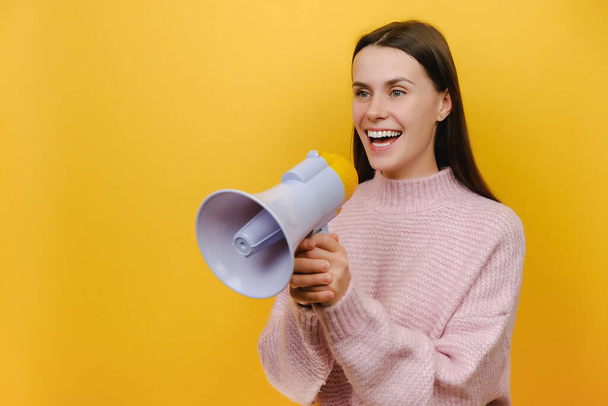 Portrait of fun expressive happy smiling student young woman scream aside shouting in megaphone, dressed in knitted sweater, isolated over yellow color background studio. People lifestyle concept - Valokuva, kuva