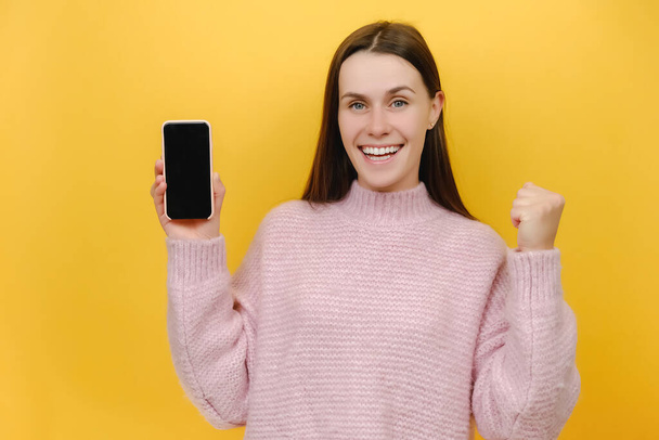 Portrait of excited overjoyed millennial female in pink sweater hold mobile cell phone with blank screen workspace area do winner gesture clenched fist, posing isolated over yellow background studio - Fotografie, Obrázek