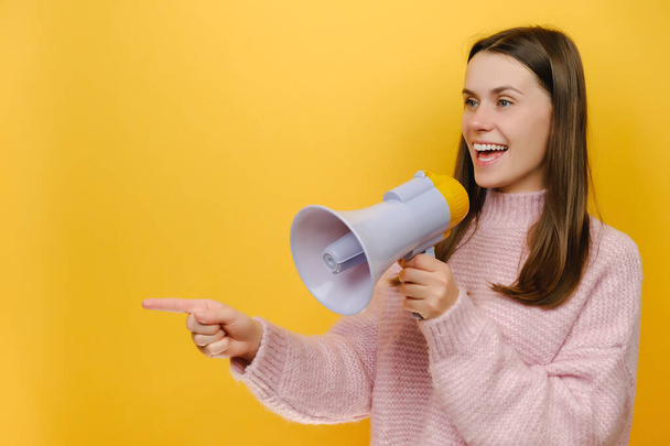Cute beautiful young caucasian woman 20s screaming hot news announces shout in megaphone point index finger aside, wears knitted sweater, posing isolated over yellow color background wall in studio - Фото, изображение