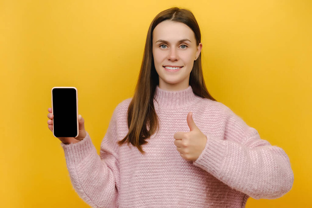 Portrait of young caucasian smiling woman 20s holding mobile cell phone with blank screen workspace area show thumb up gesture, wear pink knitted sweater, isolated on yellow color background in studio - Photo, image