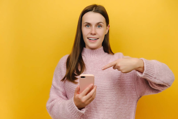 Surprised amazed young brunette woman wearing pink knitted sweater standing pointing index finger on mobile cell phone typing sms message, posing isolated over yellow color background wall in studio - Foto, afbeelding