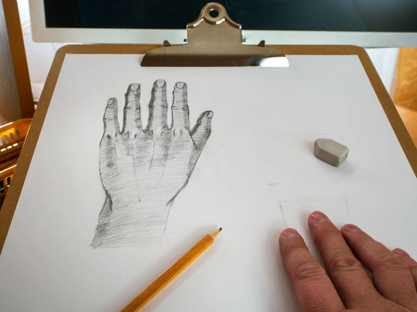A hand is drawn on a sheet of Whatman paper, there is a simple pencil and an eraser. The fingers of the hand are visible below. There is a monitor behind the picture. Online education, drawing lessons - Foto, Bild