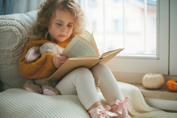 A little cute curly haired girl in knitted pullover is reading a book in front of the window. Cozy home. Warm autumn. Mood. - Φωτογραφία, εικόνα