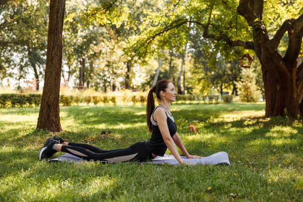 Молодая женщина в спортивной форме делает упражнения на растяжку кобры в городском парке - Фото, изображение
