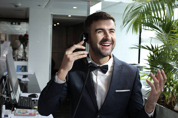 Porträt eines hübschen jungen Mannes im Smoking mit Fliege, der im luxuriösen Interieur telefoniert - Foto, Bild