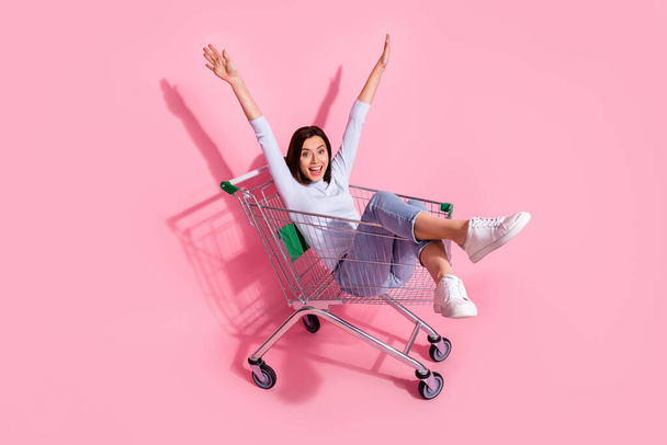 Photo of charming funny young woman wear white sweater riding shopping cart rising arms smiling isolated pink color background - Foto, afbeelding