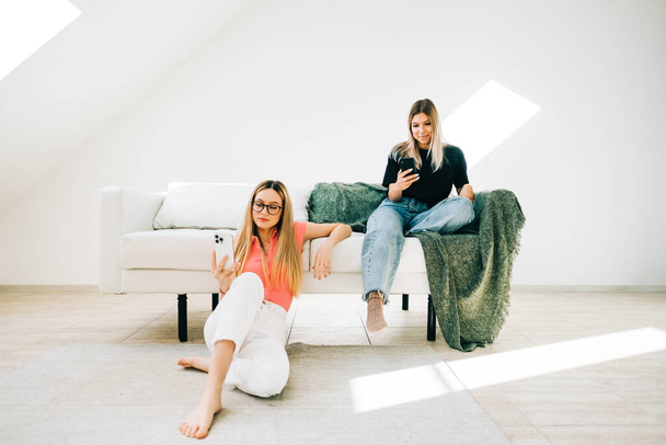 Two women friends sitting in living room on a sofa and browsing internet using mobile technology. - Photo, Image