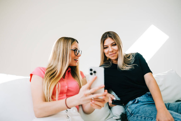 Friends heaving fun, sitting on sofa and looking on smartphone screen, two cheerful women together browsing internet. - Фото, зображення
