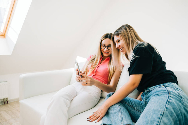 Friends heaving fun, sitting on sofa and looking on smartphone screen, two cheerful women together browsing internet. - Φωτογραφία, εικόνα