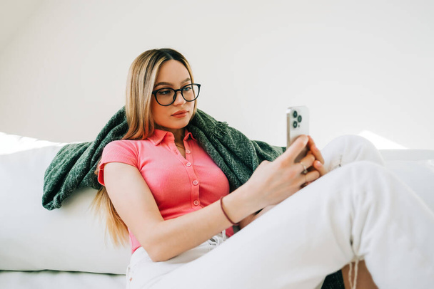 Young caucasian woman using smartphone while resting on a sofa at home.  - Фото, зображення