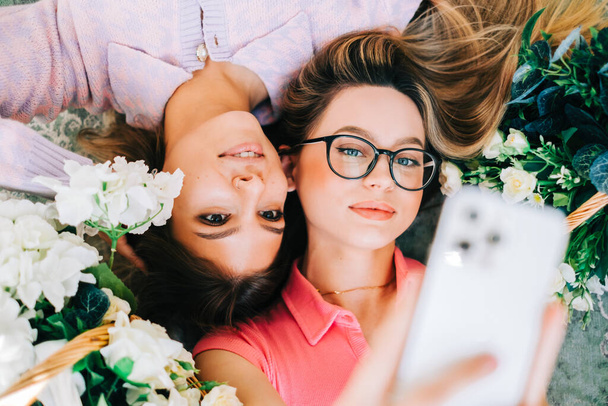 Portrait of two attractive caucasian woman lying on a floor with flowers and making selfie photo - Фото, изображение