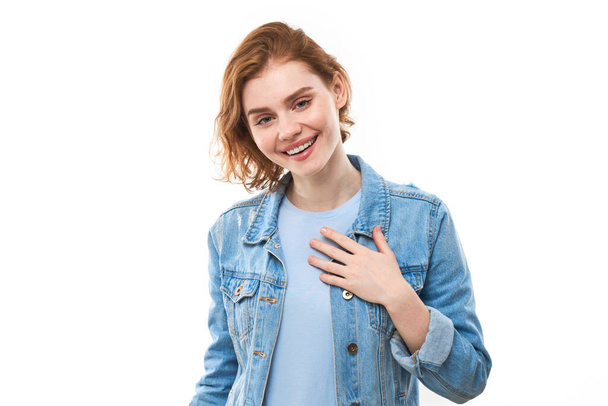 Lovely good-natured red-haired girl student in a blue denim jacket with a charming smile and a friendly expression, holding hands on her heart, wanting to show her love and sympathy. On a white background - Fotó, kép