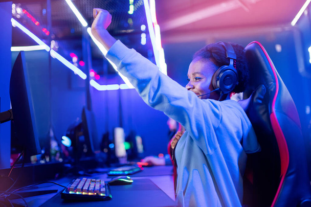 Streamer mujer joven africana se regocija en la victoria jugador profesional jugando juegos en línea ordenador con auriculares, color neón - Foto, imagen