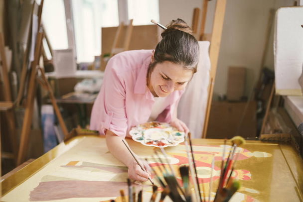 Cute woman paints on canvas in an art workshop. Artist creating picture. Art school or studio. Work with paints, brushes and easel. Hobby and leisure concept. Woman painter at workspace. - Foto, Bild