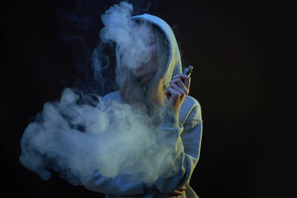 Portrait of beautiful blonde girl in gray hoodie smokes vape isolated on black studio background, cloud of steam smoke, mini hookah - Foto, immagini