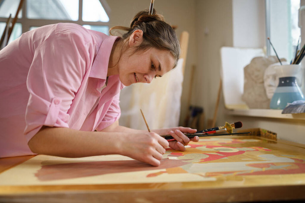 Caucasian woman artist working on a painting in bright daylight studio. Happy artist draws an art project with paints and a brush in the workshop. Hobby. Artist at work. Creative profession. - Photo, Image
