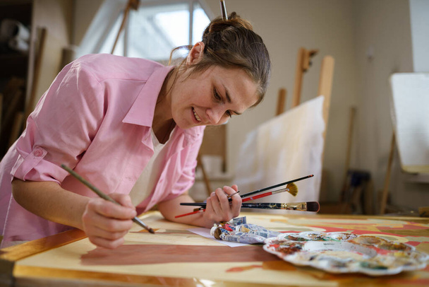 明るい昼間のスタジオで絵を描く白人女性アーティスト。ワークショップでは、絵具や筆を使ったアートプロジェクトを行います。趣味だ。仕事中のアーティスト。創造的な職業. - 写真・画像