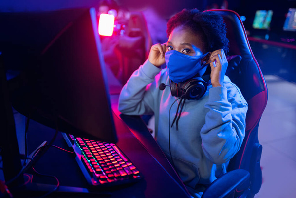 Hosting online videopeli turnauksia tietokone klubeja pandemian aikana, amerikkalainen Afrikkalainen nuori nainen yllään suojaava naamio vastaan Covid - Valokuva, kuva