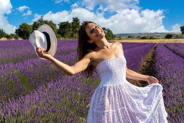 Portret van een gelukkige jonge vrouw die met haar hoed speelt in een lavendelveld. Haar witte jurk valt op tegen de paarse kleur van lavendelbloemen. - Foto, afbeelding