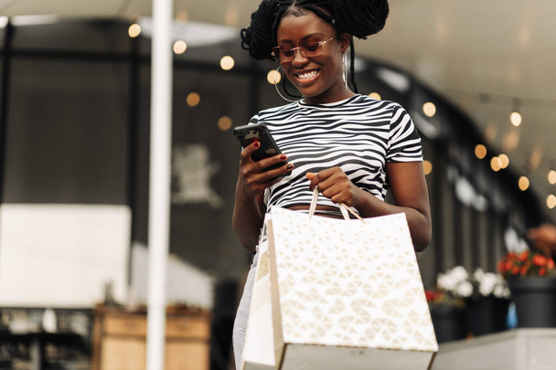Kaunis muodikas afrikkalainen amerikkalainen tyttö tilalla laukkuja, käyttäen älypuhelin ja hymyillen samalla ostoksia naisten putiikki Black Friday, ostoksia käsite, musta perjantai myynti - Valokuva, kuva