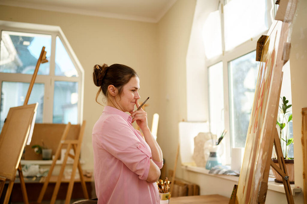 Cute woman paints on canvas in an art workshop. Artist creating picture. Art school or studio. Work with paints, brushes and easel. Hobby and leisure concept. Woman painter at workspace. - Photo, image