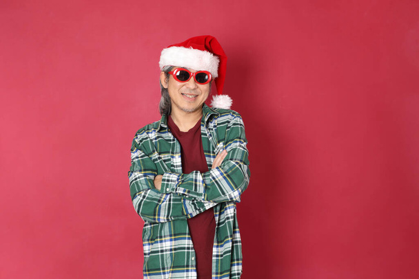 The senior Asian man wears green plaid shirt with Santa Claus hat standing on the red background. - Photo, image