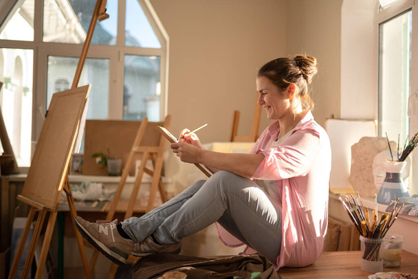 Cute woman paints on canvas in an art workshop. Artist creating picture. Art school or studio. Work with paints, brushes and easel. Hobby and leisure concept. Woman painter at workspace. - Foto, afbeelding