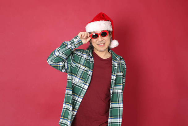 Старший азиат носит зеленую клетчатую рубашку со шляпой Санта-Клауса, стоящей на красном фоне.. - Фото, изображение